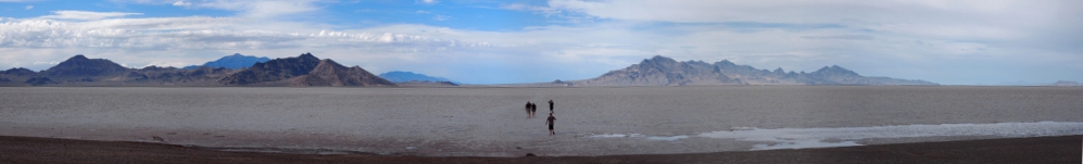 [Approximately six photos stitched together showing vast flat land with mountains in the distance. Four people (small dark splotches) are walking out onto the salt flats from the ring of dirt at the edge of the salt.]